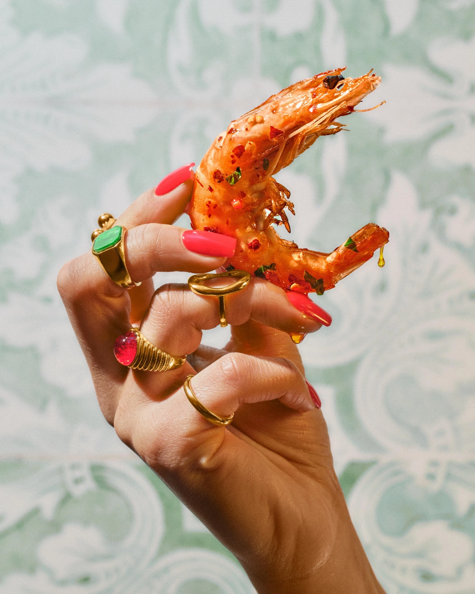 Glazed prawn held in a jewelled hand against a pale patterned background
